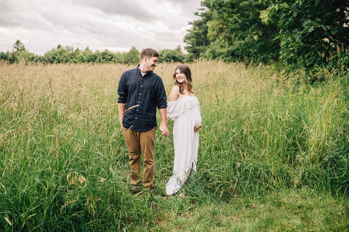 A husband and wife smiling at each other in a green field at their maternity session with Sharon Leger Photography.