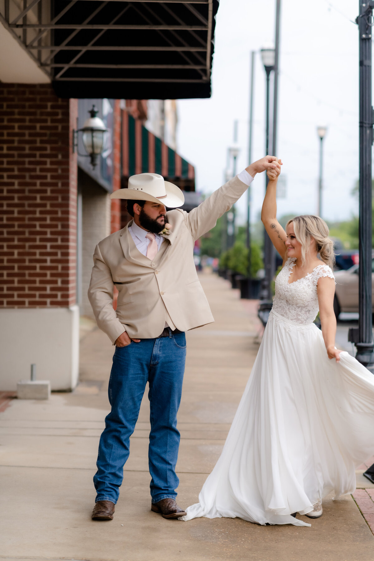groom in a beige suit coat and denim jeans twirls his bride as her lace dress flows around her
