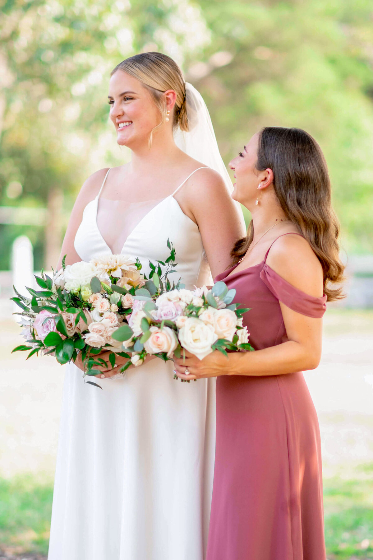 Two women outdoors, one in a white wedding dress holding a bouquet of flowers, and the other in a pink dress holding a smaller bouquet. They are smiling and standing close to each other, surrounded by greenery.