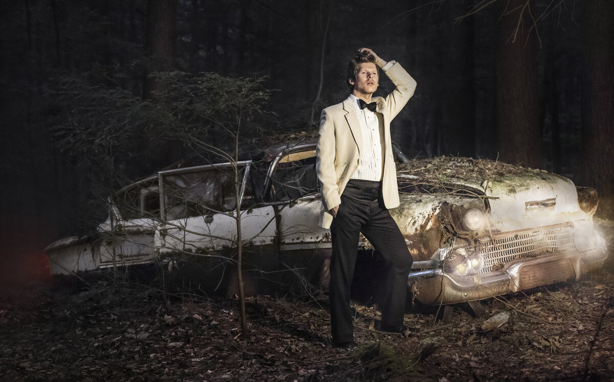 A person in a suit standing next to an abandoned car