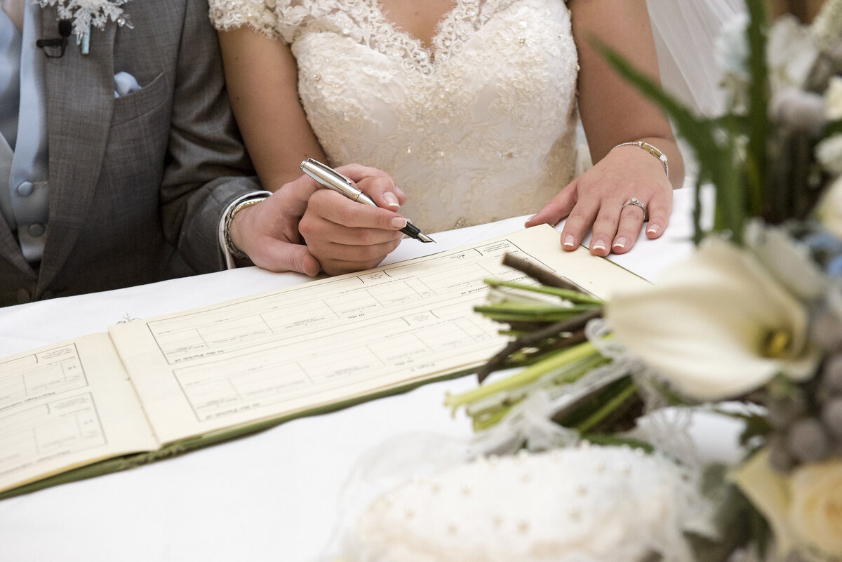 A bride signs the wedding register while her new husband holds her hand.
