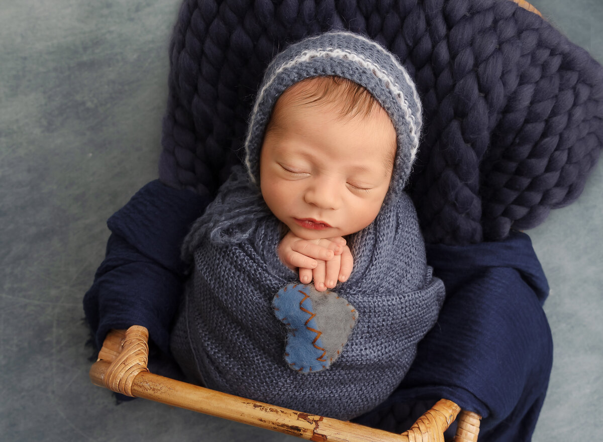 Newborn photoshoot. Baby boy swaddled in blue has his hands peeking out of the swaddle and holding a blue felt heart. Baby is placed in a basket. Captured by top Brooklyn newborn photographer Chaya Bornstein