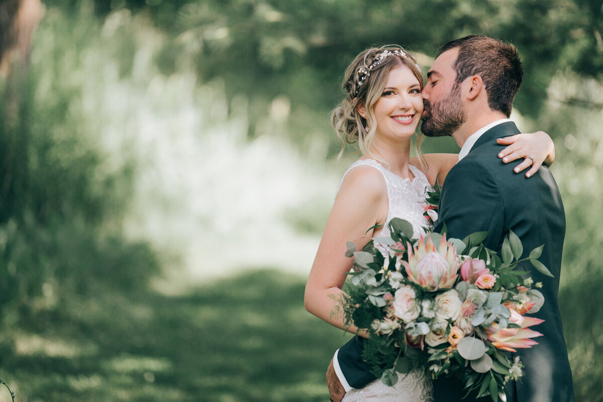 A romantic photo of a groom wearing a green suit kissing his bride while she holds a stunning, whimsical blush and green wedding bouquet