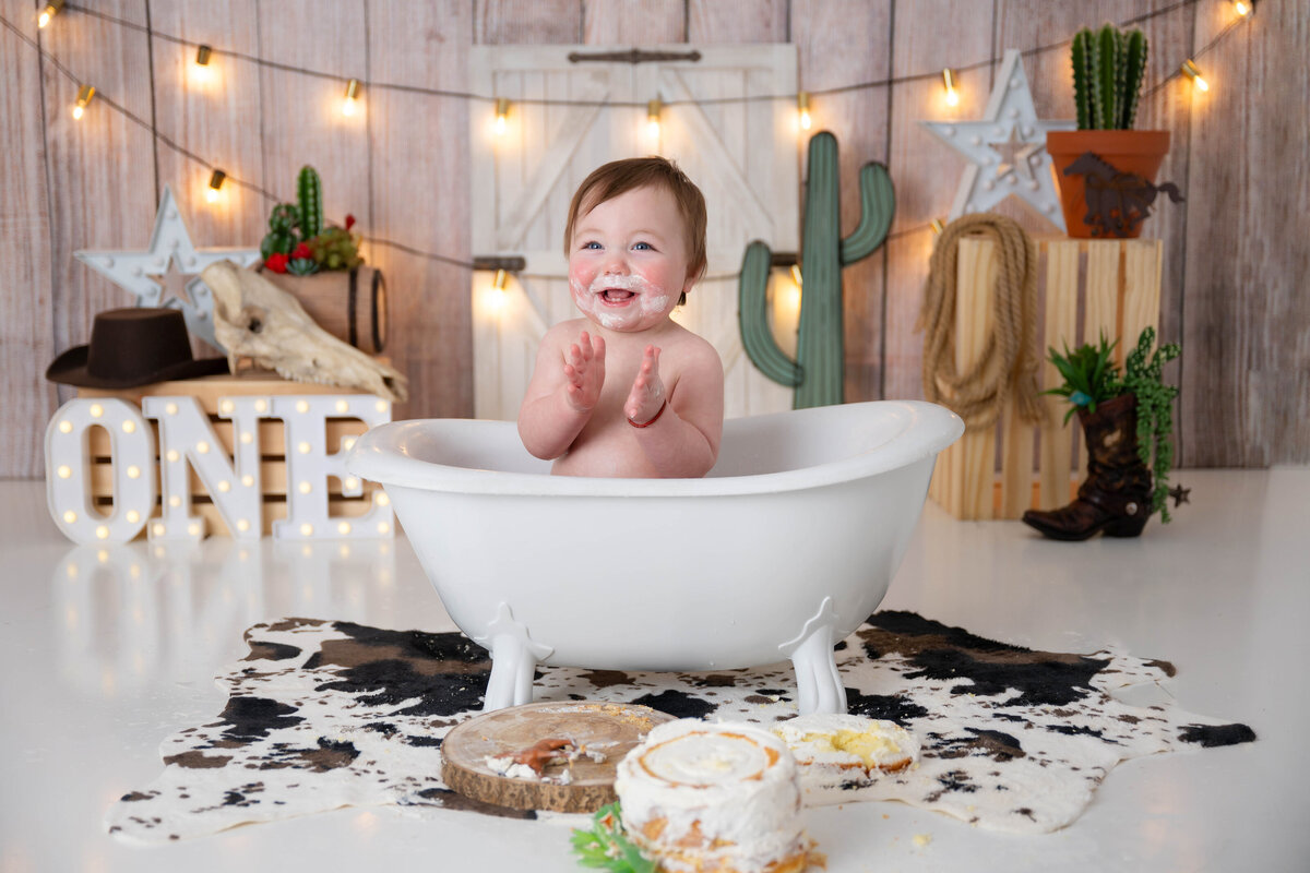 Smiling baby clapping while sitting in a mini tub, surrounded by cowboy-themed props including barrels, cow hides, and cactuses.