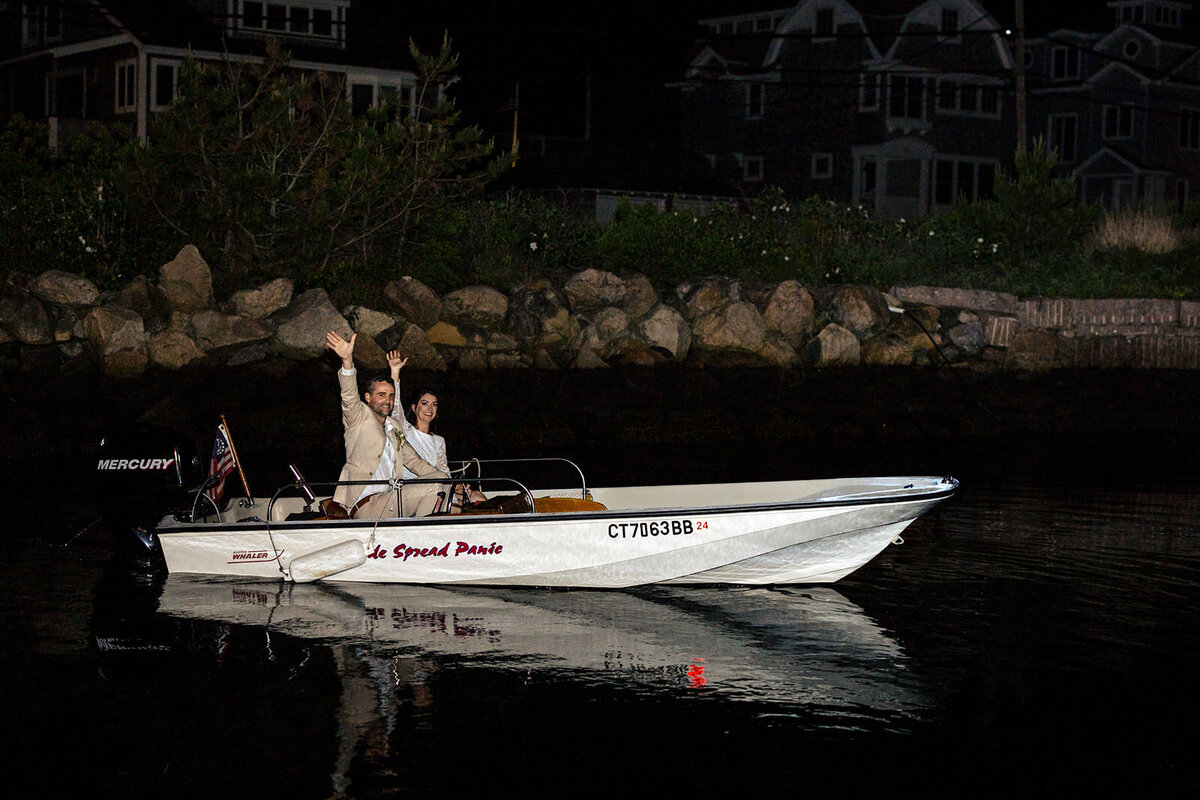 The couple waves from a speedboat named "Le Spread Panic" during a nighttime getaway, with shoreline houses in the background.