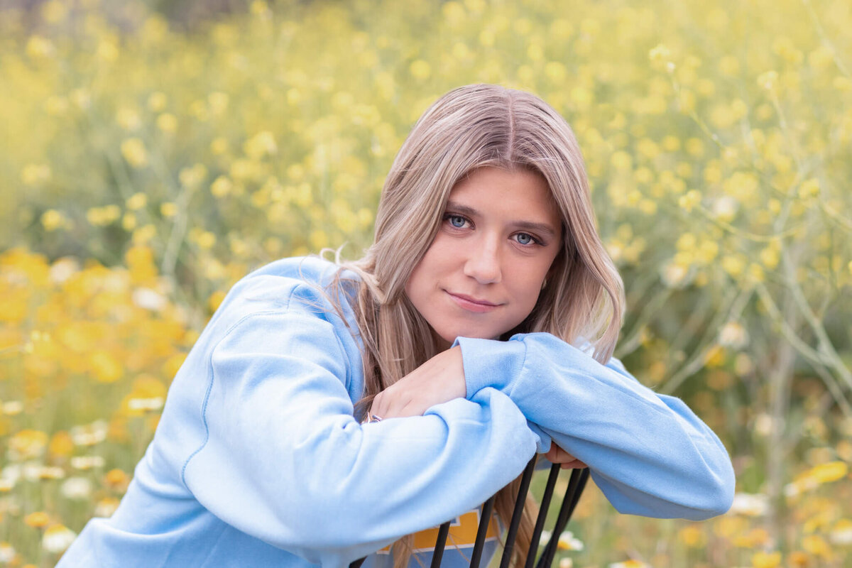 A person with long, light brown hair wearing a blue sweater leans forward on a chair in a field of yellow flowers, smiling gently at the camera.