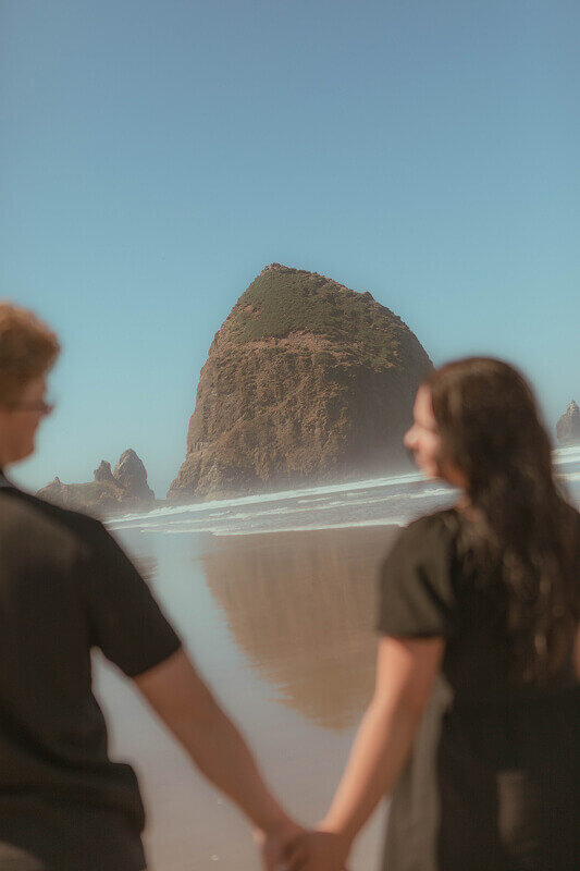 A slightly tilted photo of Haystack Rock. In the foreground is a couple holding hands who are slightly blurred.