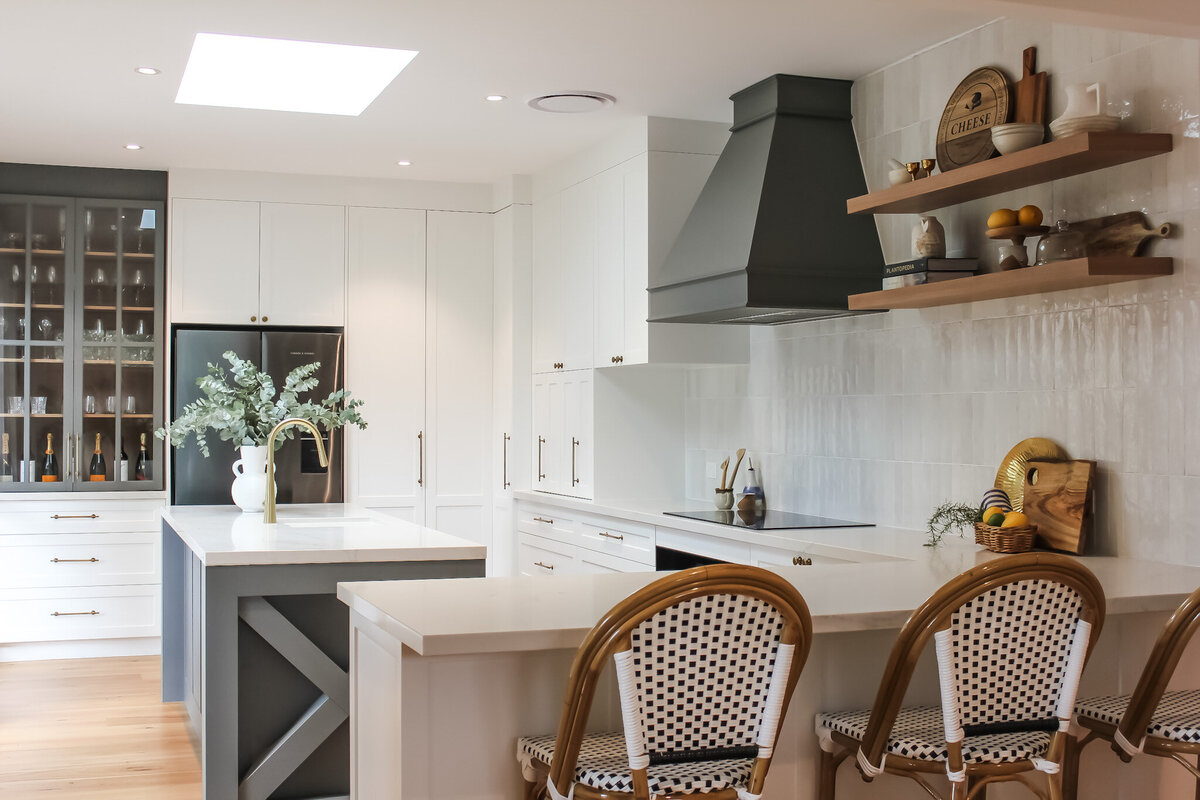 Modern kitchen with white cabinets, island, wicker chairs, and open shelving adorned with decor items, crafted by a bespoke interior design expert in Newcastle NSW.