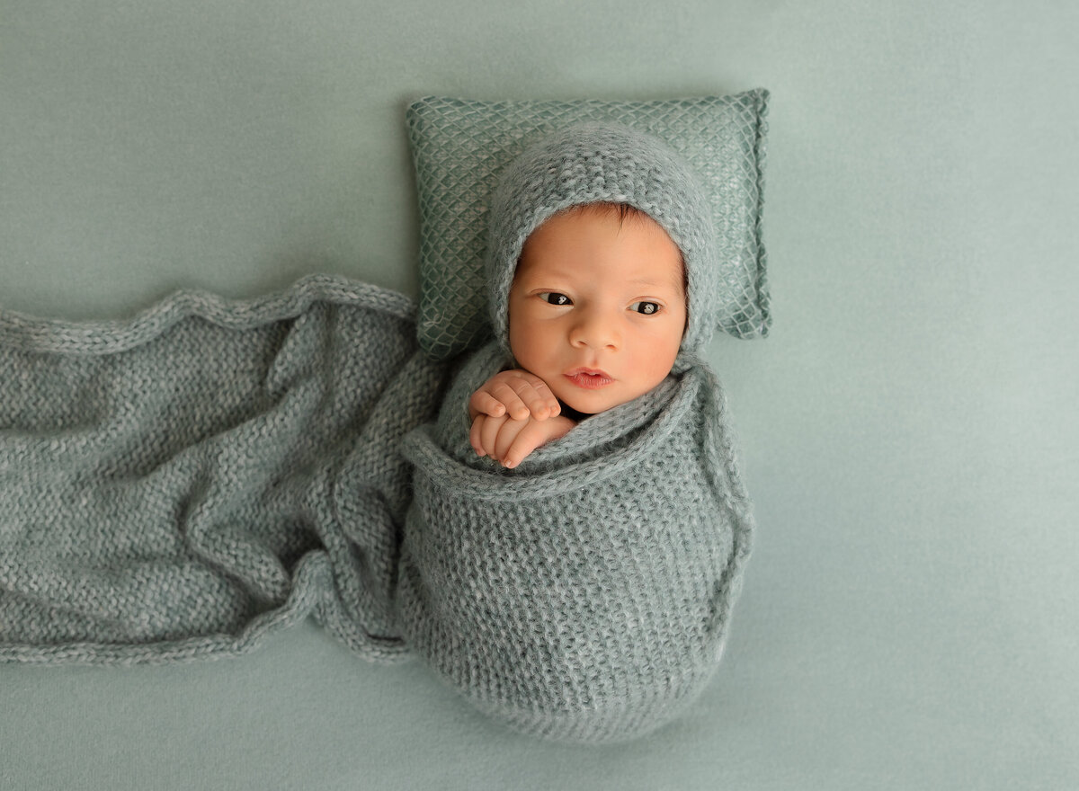 Brooklyn newborn photoshoot. Alert baby boy is swaddled in green knit with his hands peeking out of the swaddle. He is wearing a matching bonnet and laying atop of a matching knit pillow and fabric. Captured by top Brooklyn newborn photographer Chaya Bornstein