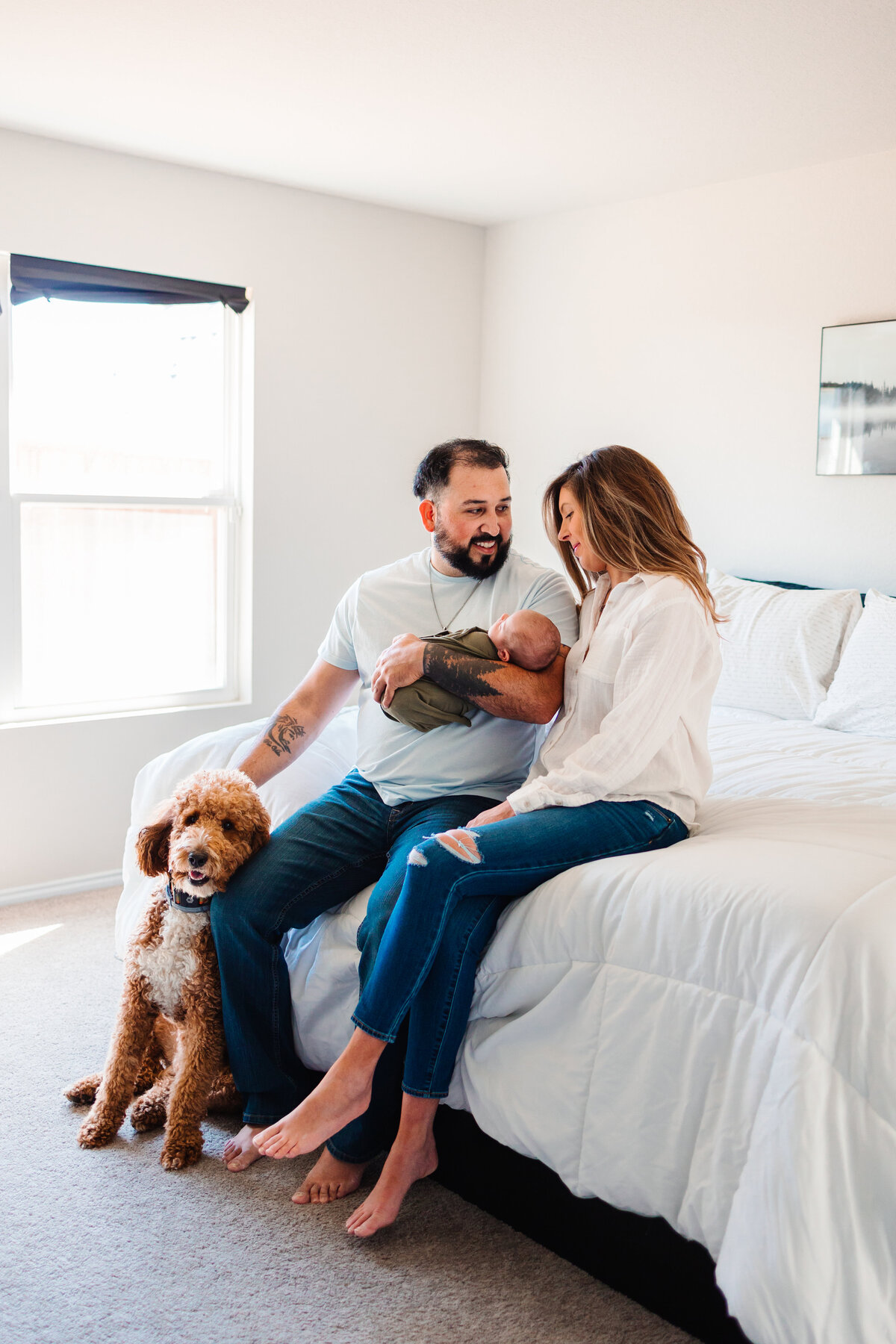 The beauty of family moments with Albuquerque maternity photography. This heartwarming image features a family with their newborn baby sitting on a white bed, all dressed in white t-shirts and blue jeans. The father holds a brown puppy, adding an extra touch of love and joy to the scene.