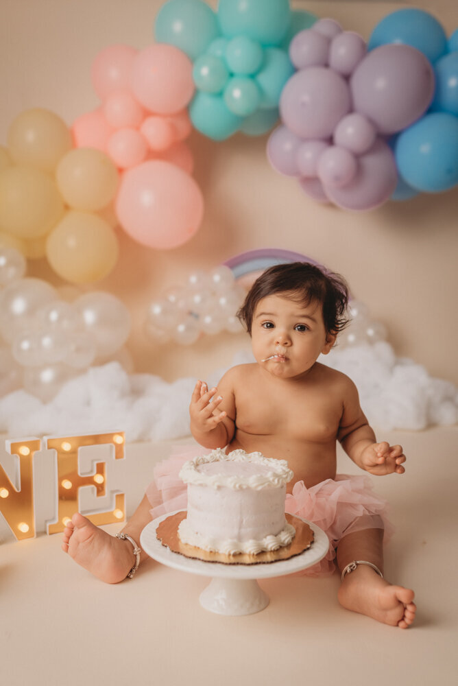 Photo shoot at Atlanta, Georgia cake smash photographer studio with one year old little girl in pink tutu smashing a cake with rainbow pastel balloon garland behind her with white fluffy clouds.