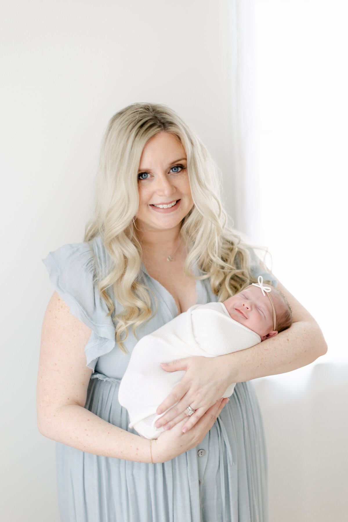 beautiful blonde mom wearing a baby blue dress holding her newborn baby girl during her newborn session photographed by Philadelphia Newborn Photographer Tara Federico