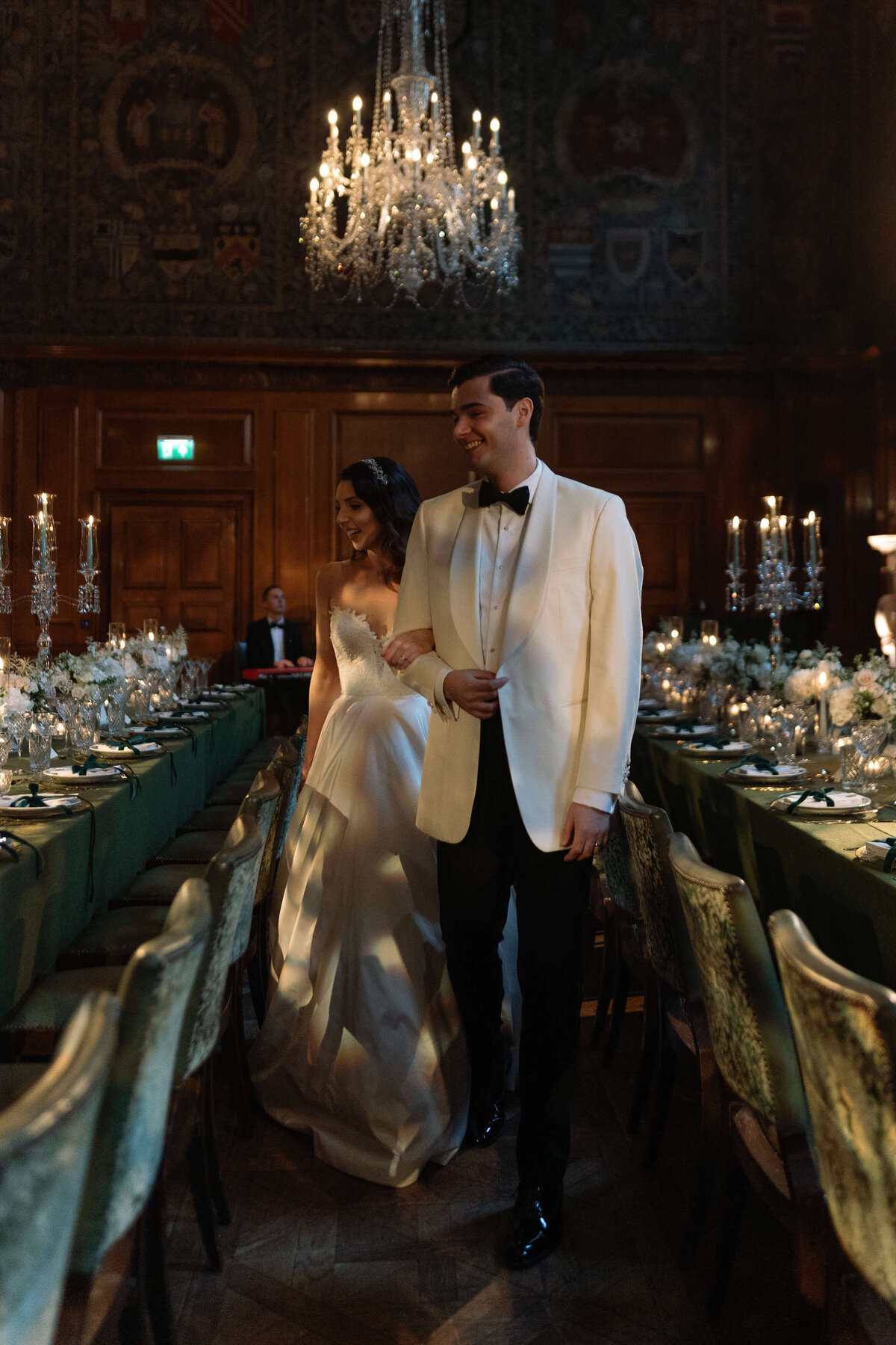 bride and groom walk between their green and gold wedding dinner tables at the ned london for their luxury city wedding as a chandelier glows above them