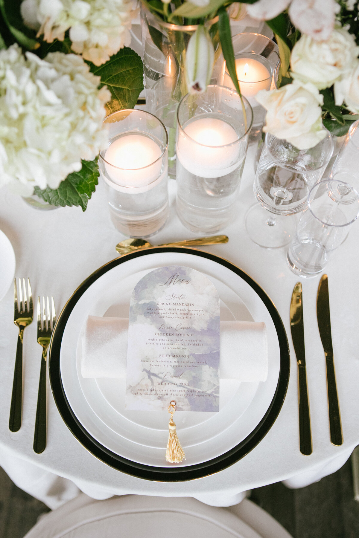 a table with a white table cloth and white flowers featuring a dinner setting with an arched menu with a gold tassel