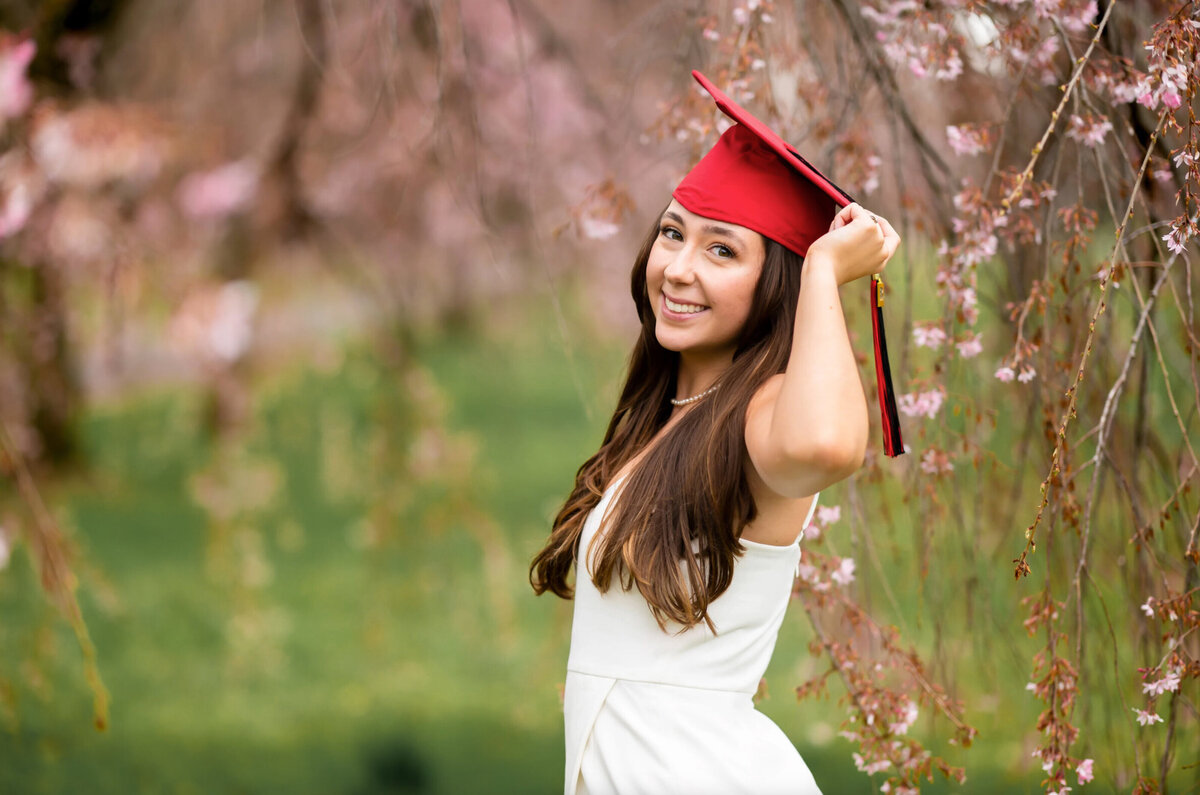 College senior, wearing a white dress and a red graduation cap, standing in front of cherry blossoms