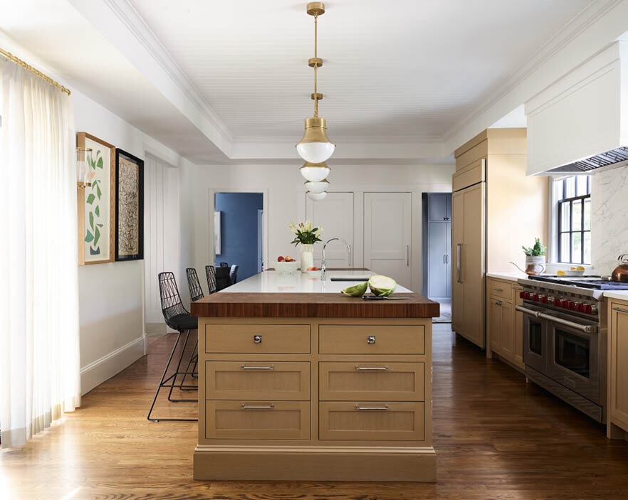 Get an overall view of the custom kitchen in a historic Hingham home, highlighting the island's edge and three equal-sized doors at the far end, leading to closets, a laundry room, and a powder room.