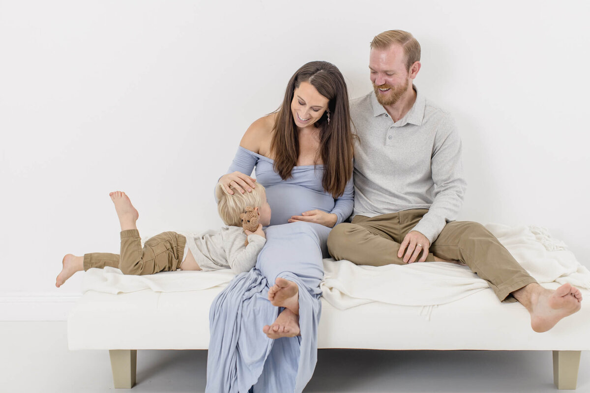 A pregnant woman in a blue dress sits on a bed with a man and a young child. The child is playfully interacting with her, while the man smiles at them. The setting is bright and minimalistic.