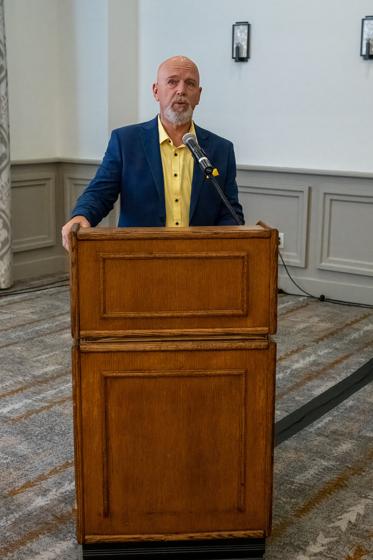 Man standing behind a wooden podium with a microphone, wearing a blue blazer and yellow shirt, photographed by Vyrl Photo for professional business presentation branding.