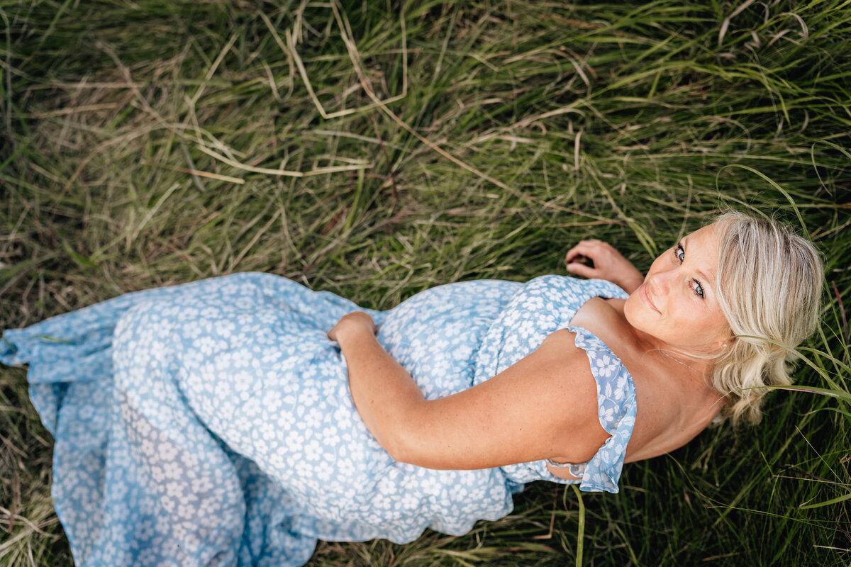 A pregnant woman lies on her side in a grassy field, looking up at the camera while wearing a light blue floral dress.