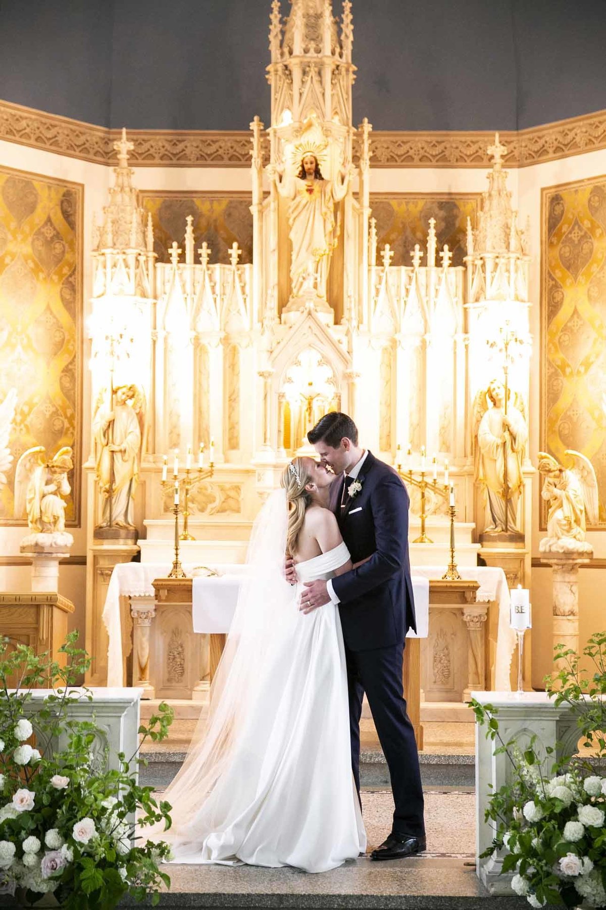 bride and groom kissing in front of altar at Villa Academy chapel with white and green flower arrangements in front