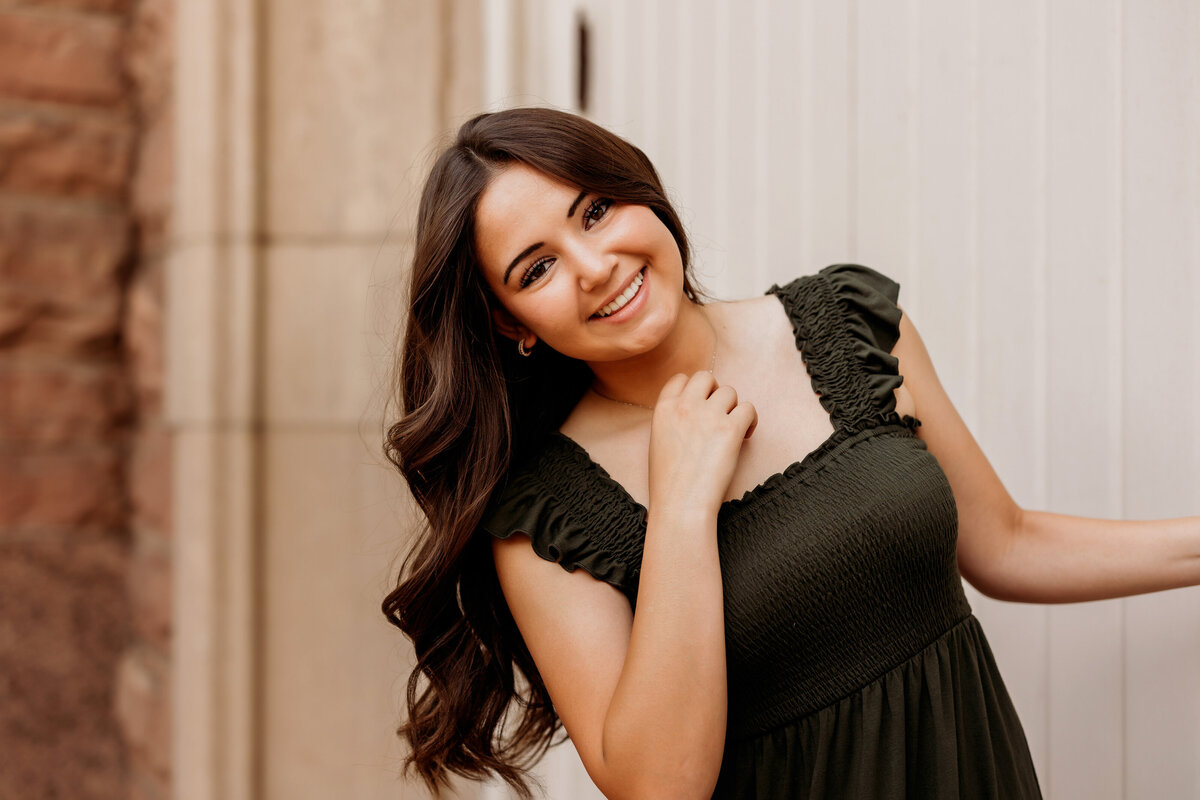 Senior girl with long brown curled hair hangs off white barn door handle during her senior photos.