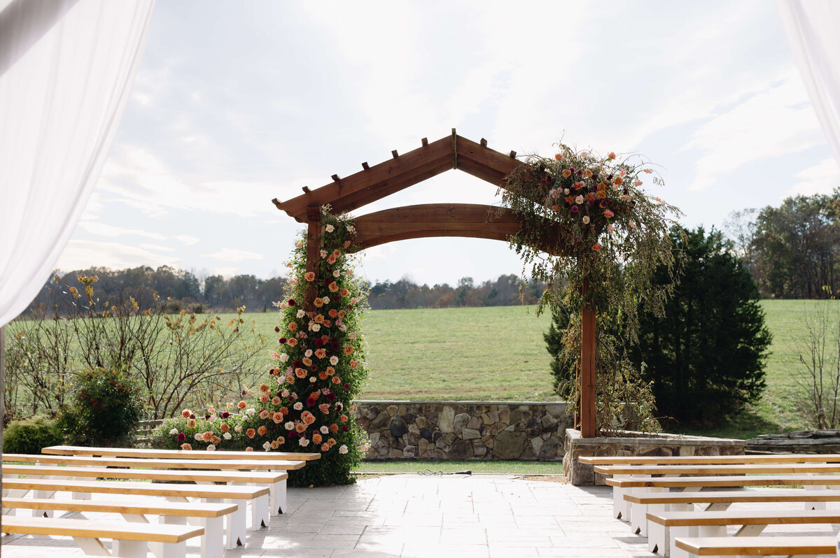 Charlottesville wedding venues ceremony space outside with a large wedding arch covered in floarl arrangements with a green hill and tree line in the distance with wooden benches leading to the arch photographed by Virginia wedding photographer