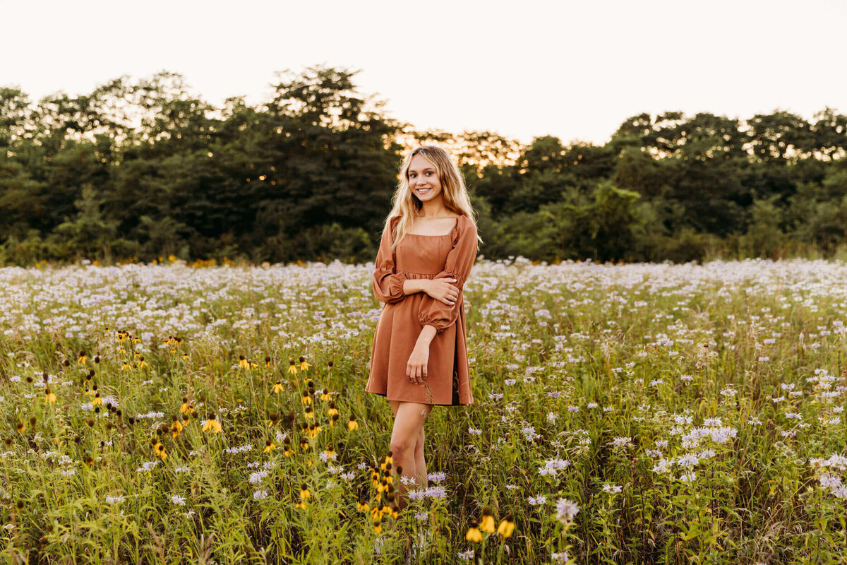 blonde senior girl standing in a field of wildflowers at sunset in an orange dress