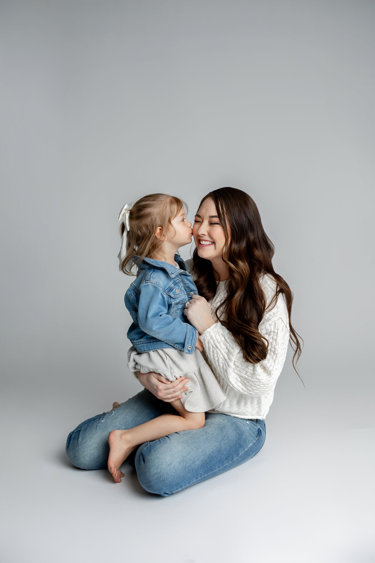 Mother cuddling daughter in her lap, both smiling warmly in studio setting. Photographed by Yucaipa family photographer Kaitlyn Dawn Photography.