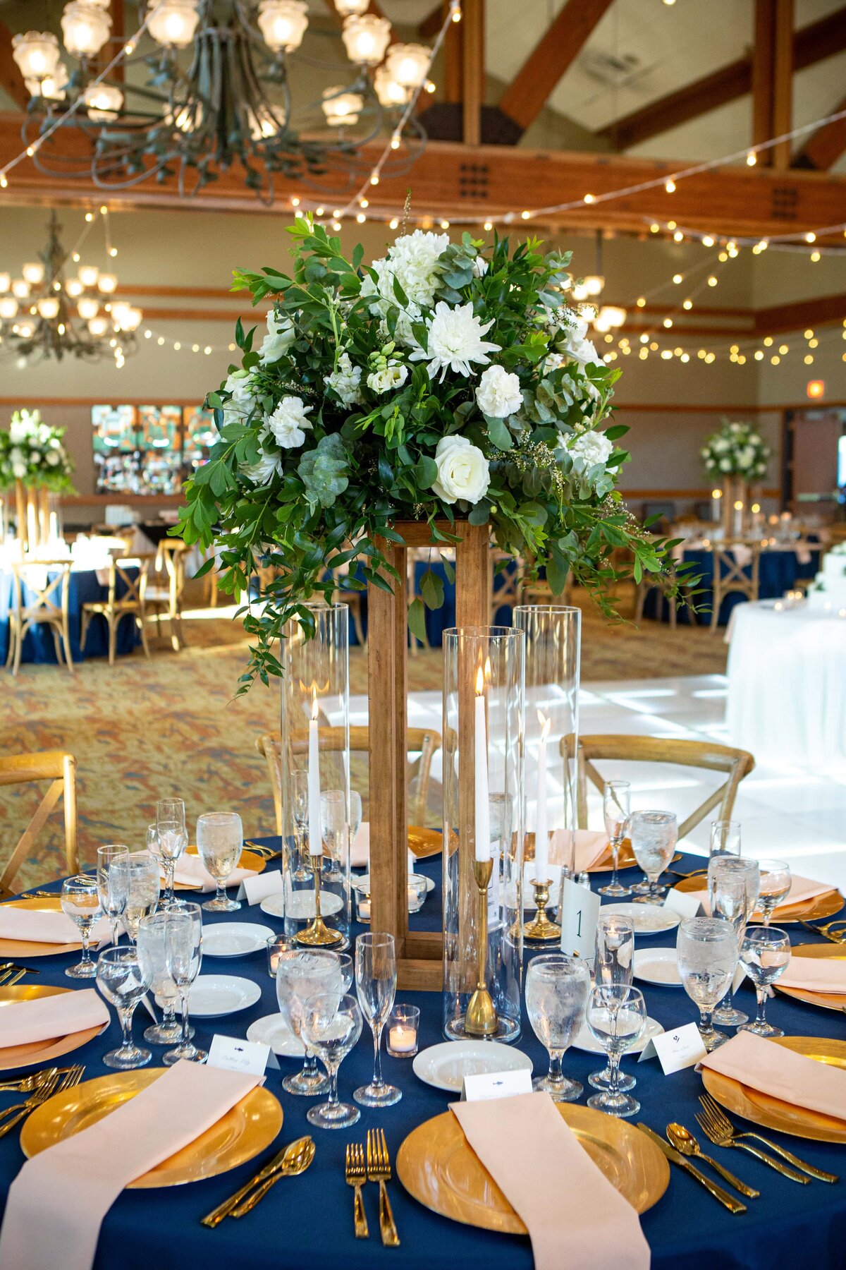 Elegant wedding reception table setting featuring blue tablecloths, gold chargers, and a tall floral centerpiece in a well-lit venue with string lights, masterfully arranged by an Illinois wedding planner.