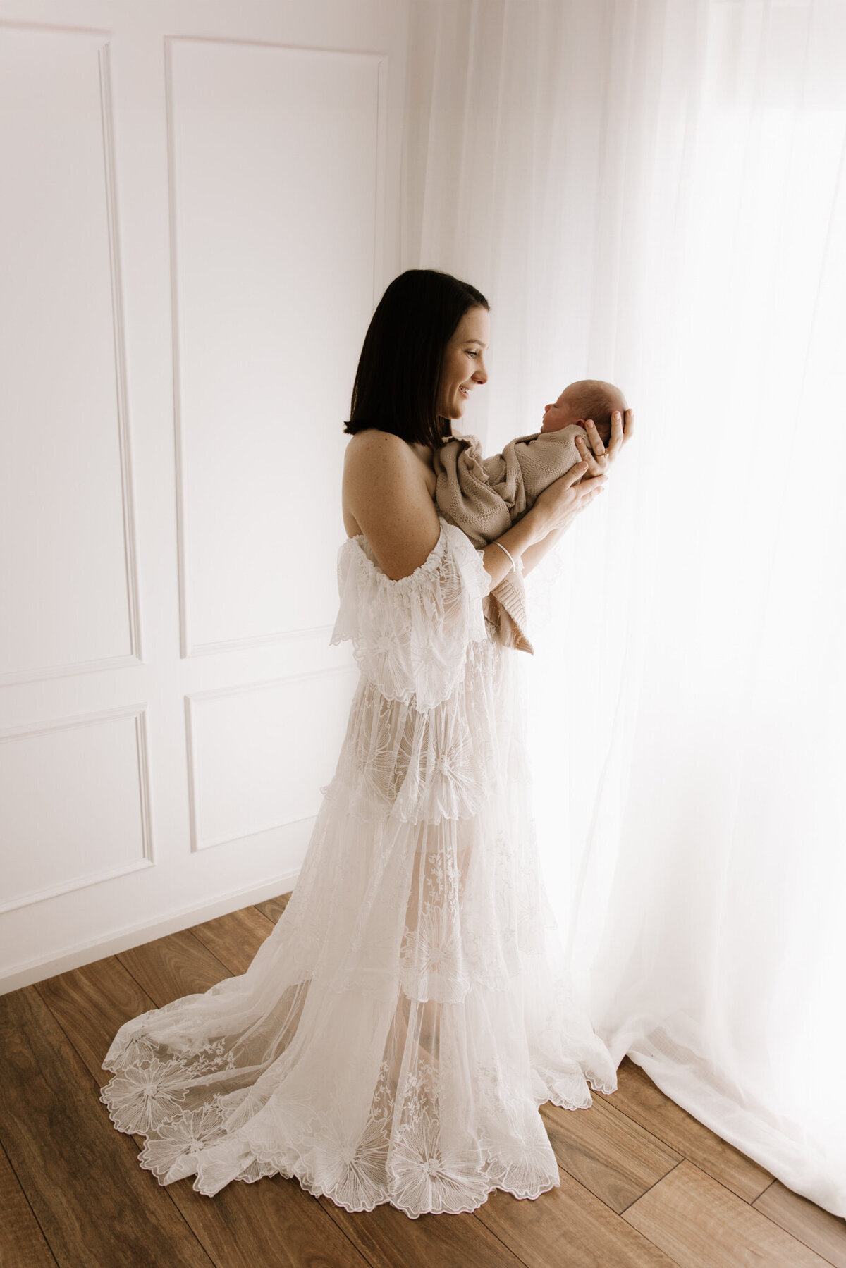 Full-length portrait of a mother in a white lace gown holding her newborn baby in a softly lit studio, captured by a Southwest Sydney newborn photographer.