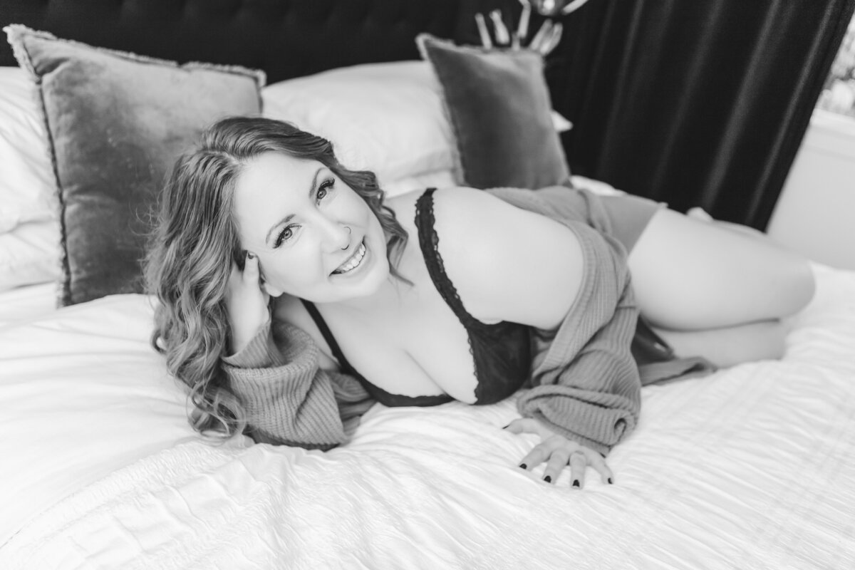 A woman lounging playfully on a bed, wearing black lingerie and a mustard cardigan, with a warm and inviting smile.