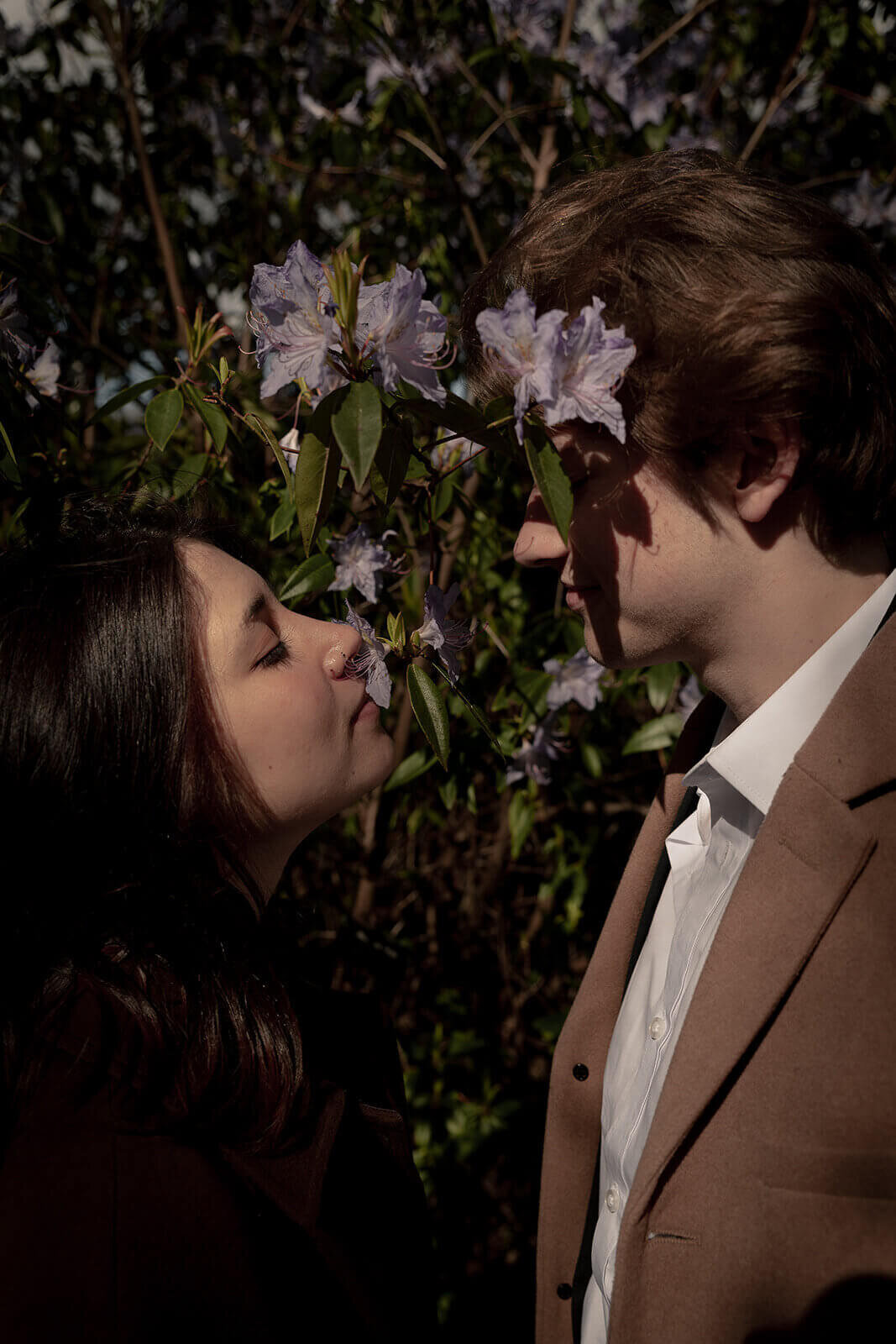 A couple smell the blossom, the shadows are across the mans face, they look lovingly at each other.