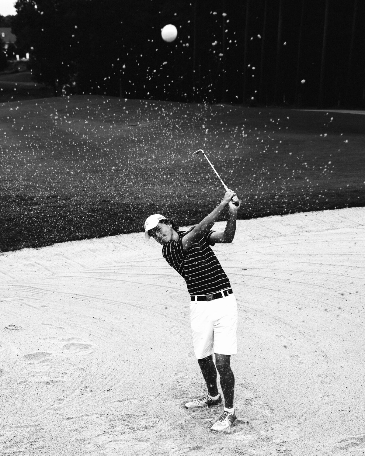 Black and white image of Garrett Risner swinging golf club in sand trap below camera with ball flying over photographer and sand flying through the air. Captured at The Club at 12 Oaks Holly Springs, NC in Spring 2020.