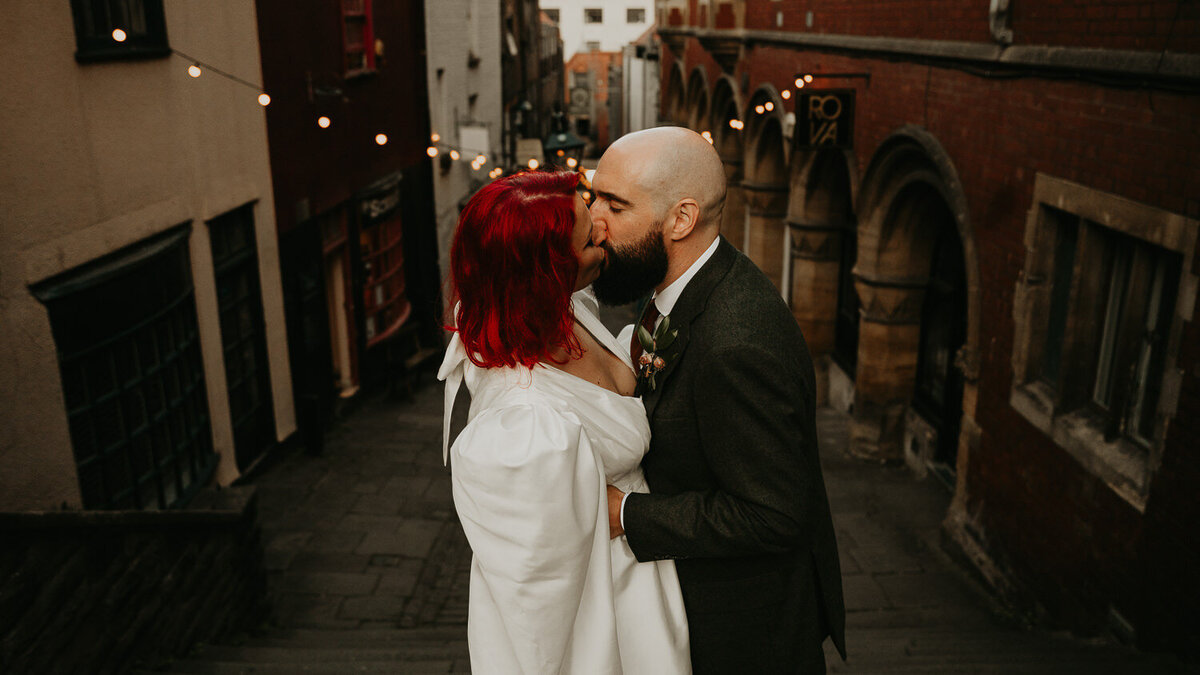 Bride wearing a short wedding dress and red hair kisses her groom on the Christmas Steps in Bristol on their wedding day. 