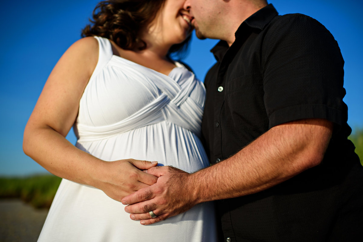 A husband and wife place their hands on her pregnant belly on the beach.