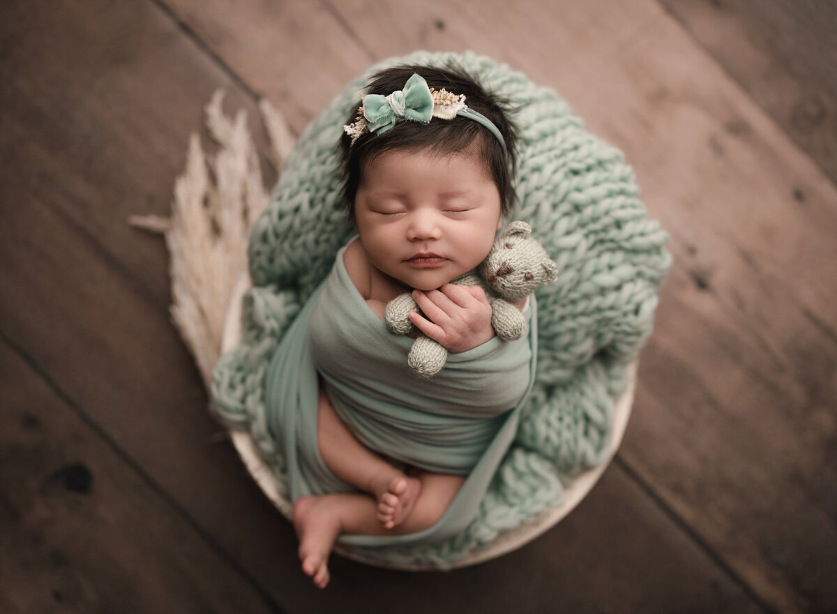 Aerial image of Riverside, CA newborn photoshoot. Baby girl is wrapped in a sage green swaddle with her legs out of the wrap and folded atop of her  Her hands are peeking out. She is holding a small felt teddy bear. Captured by Riverside's best newborn photographer Bonny Lynn Photography.
