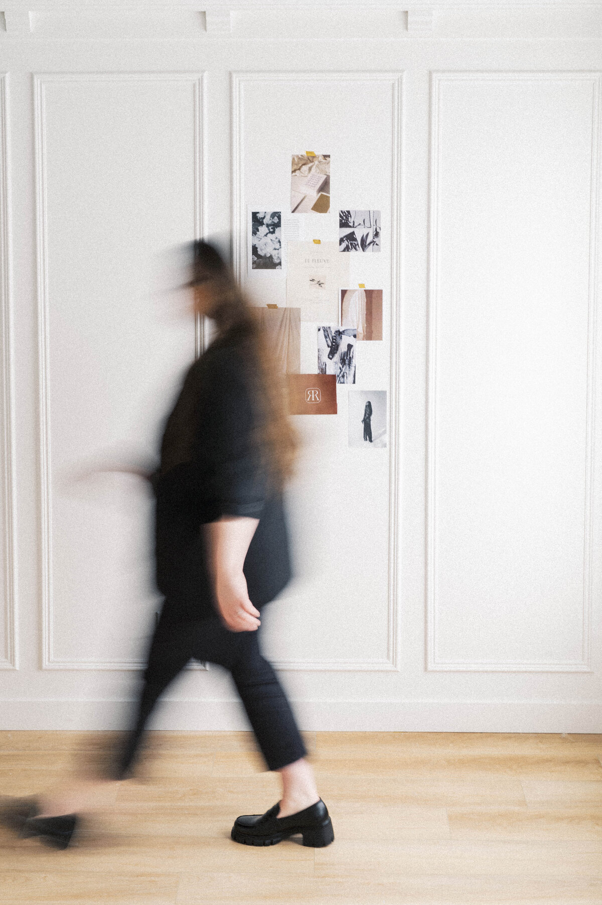 Blurry woman wearing black walking by a white wall with moodboard