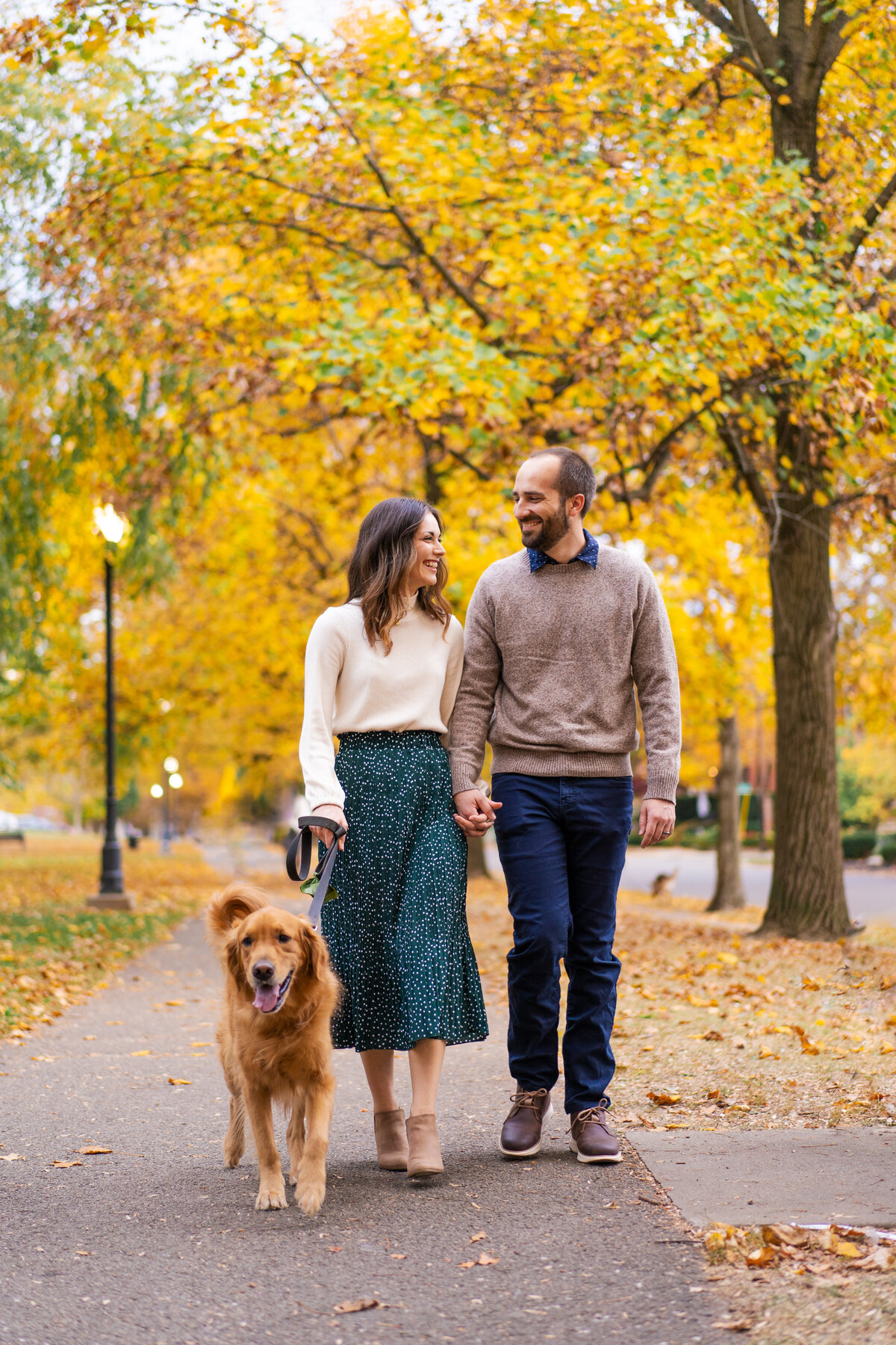 A couple and their Golden Retriever walk together and smile  amongst fall foliage at Schiller Park.