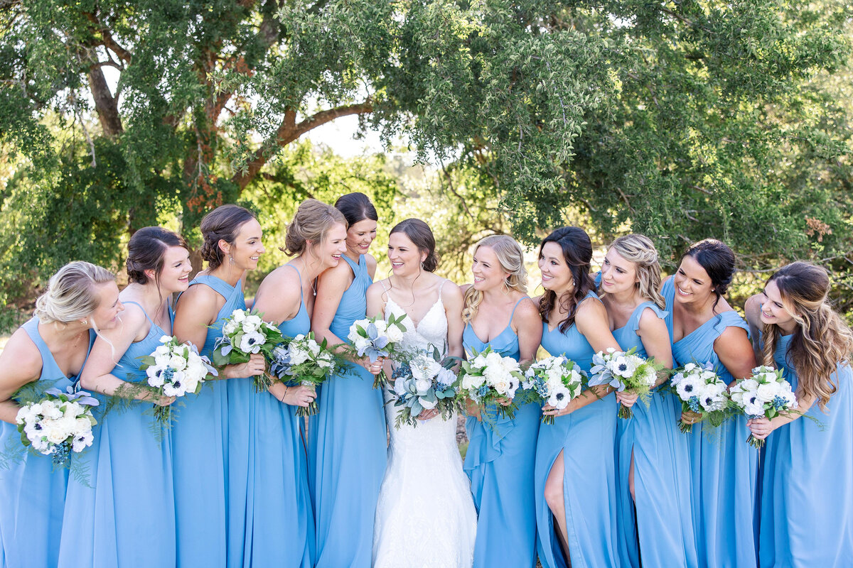 bride and 10 bridesmaids in blue laugh with each other at Morgan Creek Barn wedding in Dripping Springs Texas
