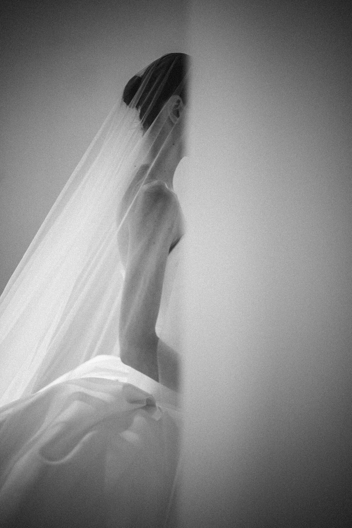 Black and white image of a figure in a structured gown and flowing veil, partially obscured by a soft foreground element—an abstract, editorial-style wedding photo taken at the New York Public Library, capturing elegance through light and form.