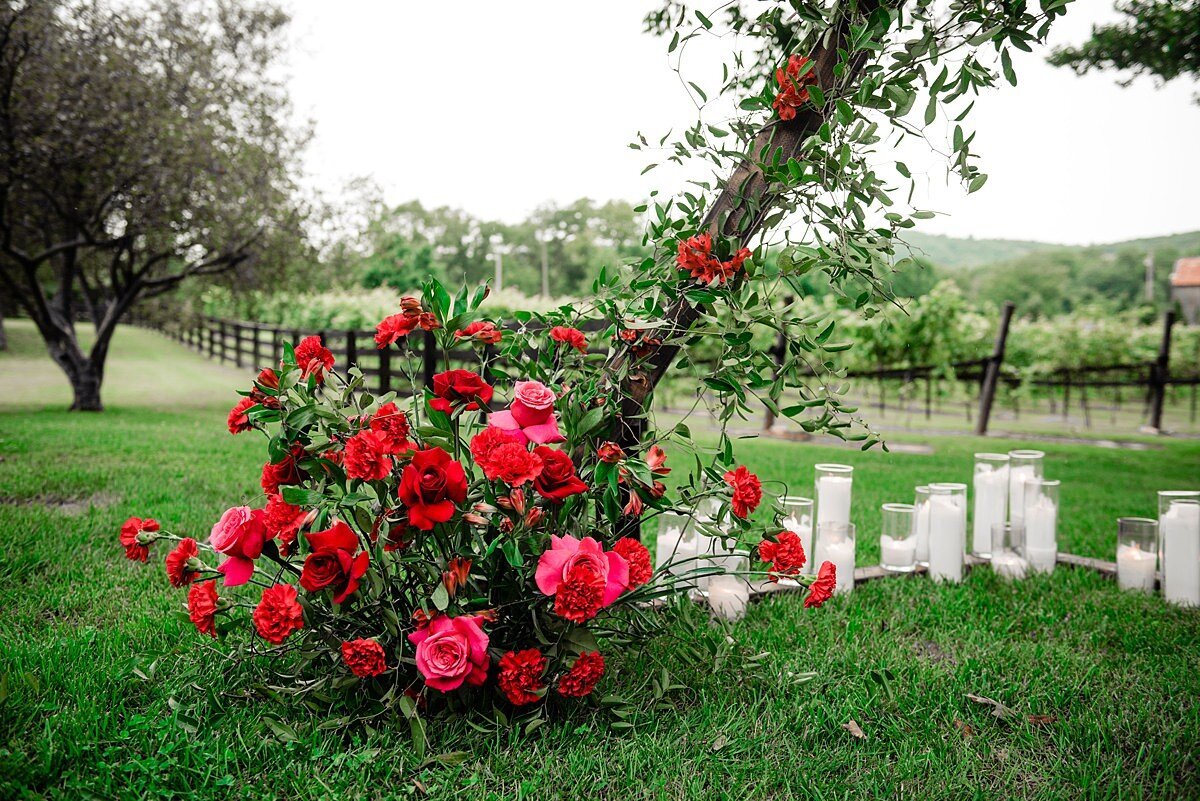 Large centerpiece of red roses, red carnations and greenery at the base of a ceremony arbor with lush smilax vine  wound up the side of the arbor accented with pink and red roses. The ceremony arbor is accented with a long row of white pillar candles in glass cylinders looking out over the trees and vines at Arrington Vineyards.