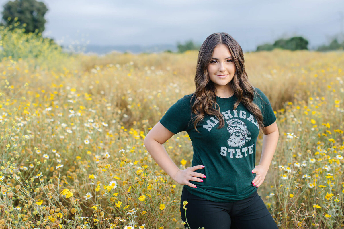 A person with long hair stands in a field of yellow and white wildflowers, wearing a green Michigan State t-shirt and black pants, smiling at the camera. The sky is overcast.