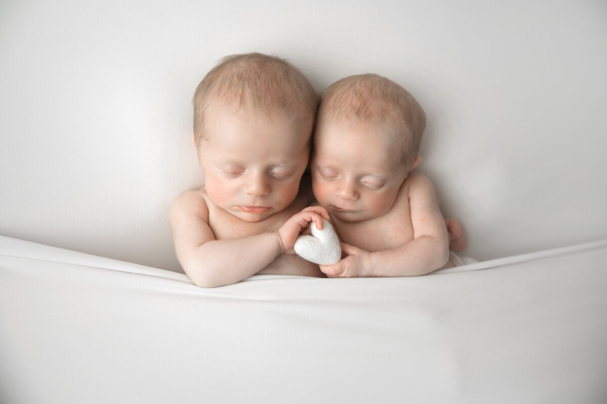 Baby girl twins hold a heart between them, cuddling together.