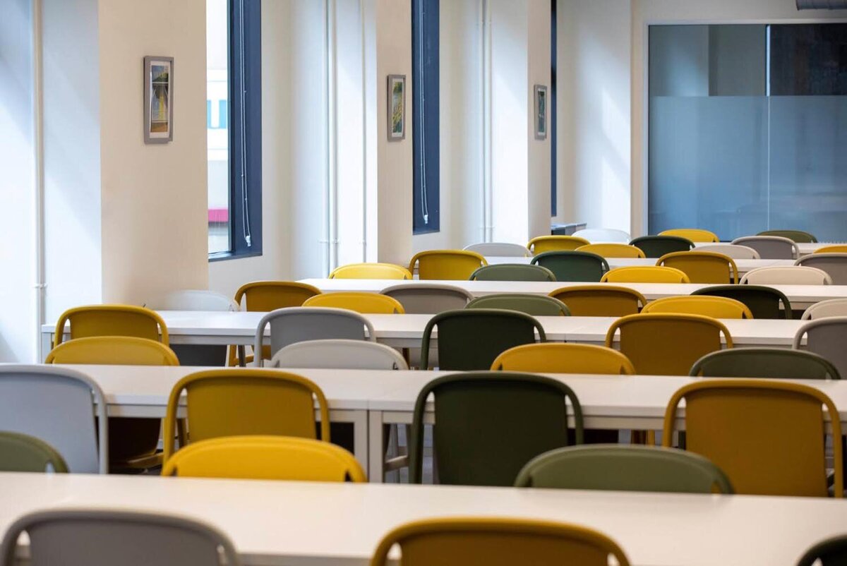 Large, brightly lit training room with rows of white tables and yellow, gray, and green chairs facing tall windows.