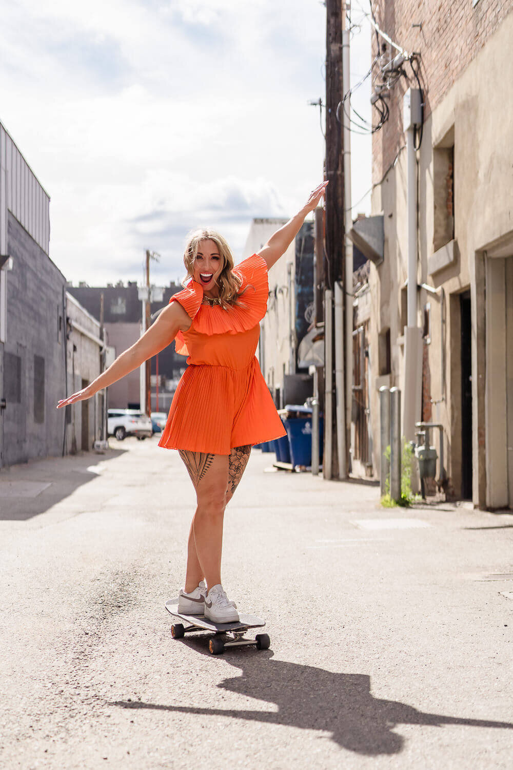 Entrepreneur in bright orange romper skateboarding with arms outstretched for balance in Kelowna alley.