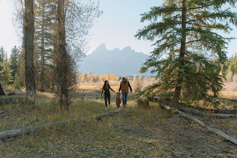 A married couple walk towards the camera with their daughter between them. Behind them are stunning views of the Grand Tetons in eastern Idaho.