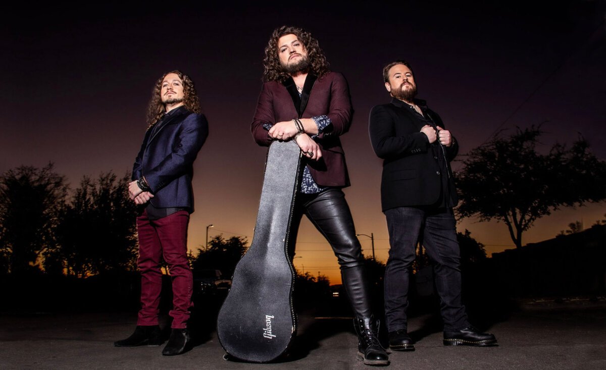 Musical trio portrait Radiofix standing roadside middle member leaning against guitar case sunset background