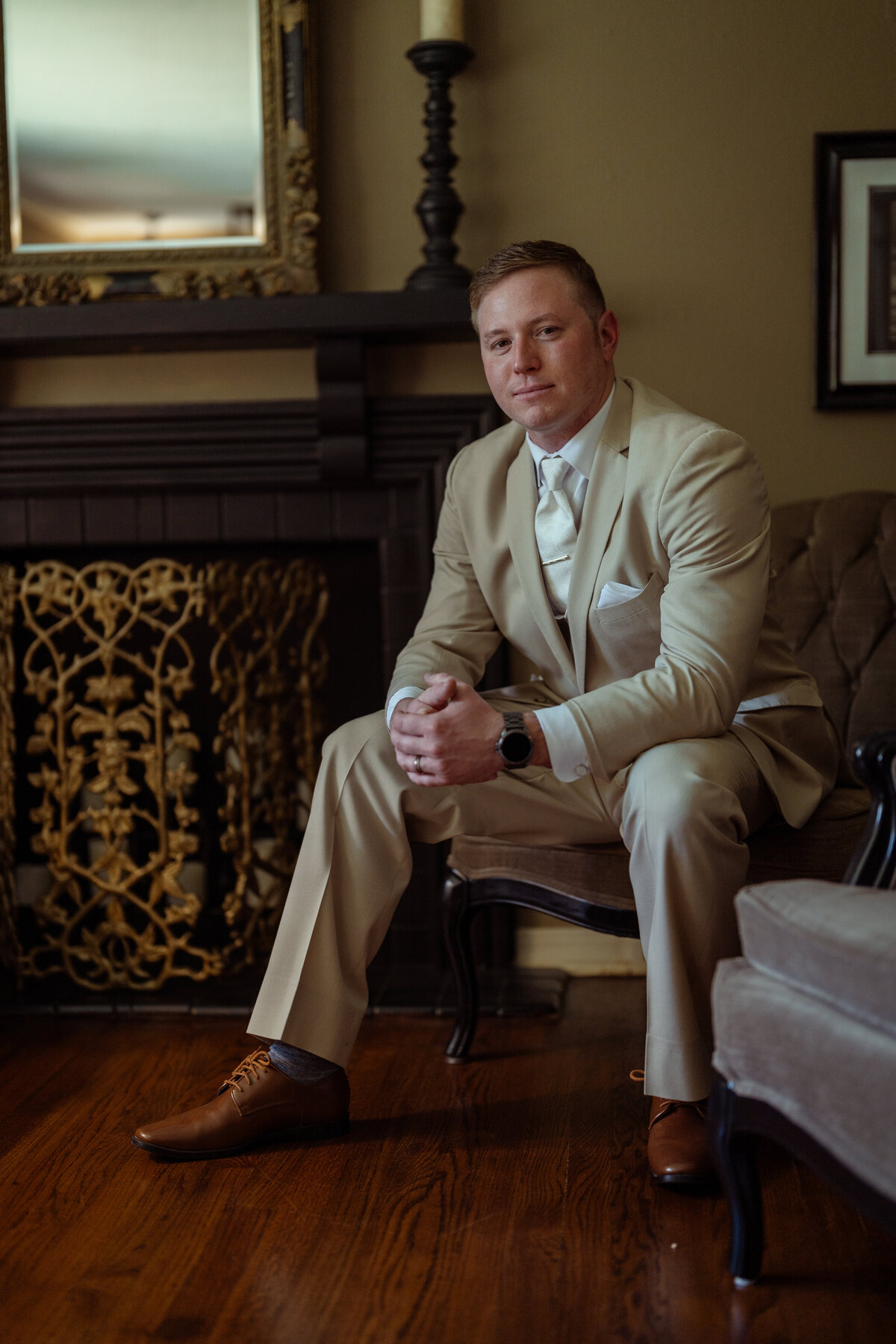 Elegant portrait of a groom sitting calmly in a luxurious setting moments before his wedding ceremony in Fort Worth. Dressed in a beige suit with matching tie and polished brown shoes, the groom exudes sophistication and confidence, captured beautifully by a luxury wedding photographer.