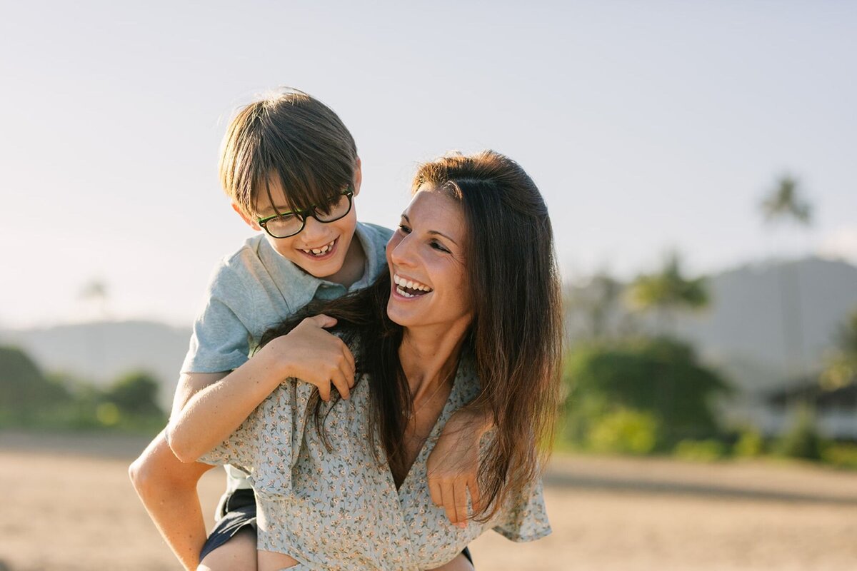 A mother gives her son a piggyback ride, as both of them are laughing on the beach.