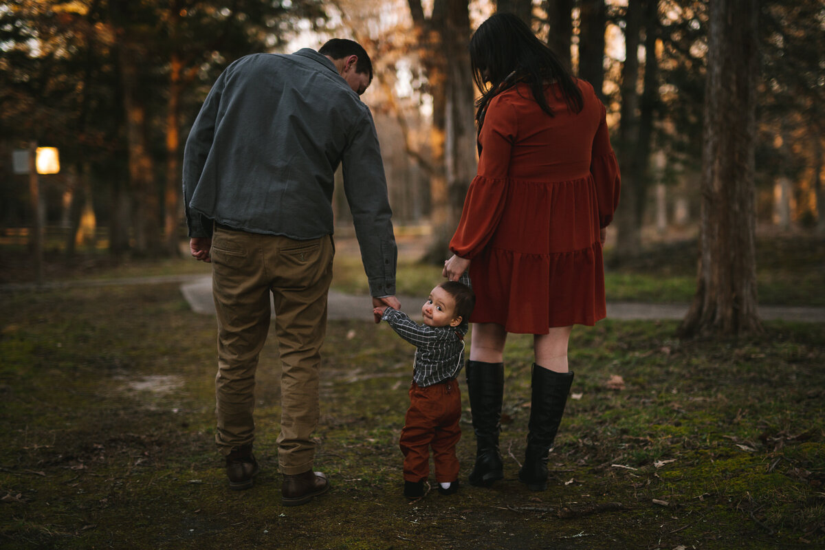 Family walks through the park, mother in a burnt orange autumn dress with riding boots, father in corduroys and a button down shirt with brown shoes as their child holds their hands looking back at the camera.